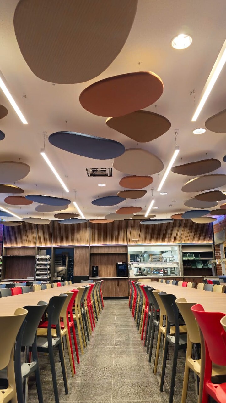 Mensa des Mediacampus Frankfurt. Zentraler Gang, links und rechts sind St&uuml;hle an Tische rangeschoben. An der Decke h&auml;ngen Akoustik-Elemente in organischen Formen. Das Licht ist an und leuchtet die gesamte Mensa hell aus.